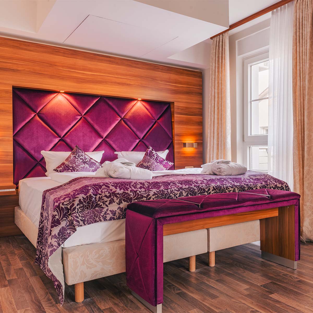 Doppelbett Suite in der Ferienwohnung in Binz auf Rügen mit violettem Kopfteil und eleganter Bettbank vor hellem Fenster.