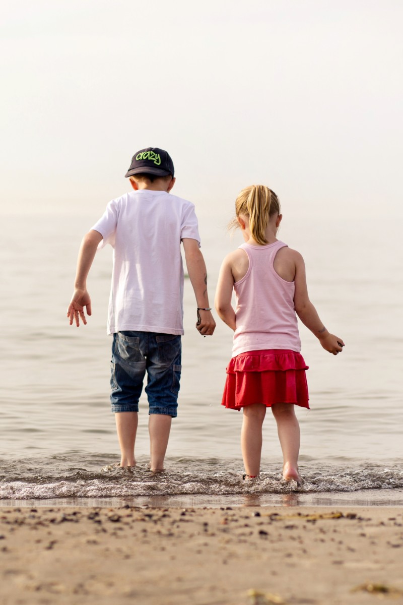 A boy and a girl are standing in the water on the beach at dusk, looking out at the sea.