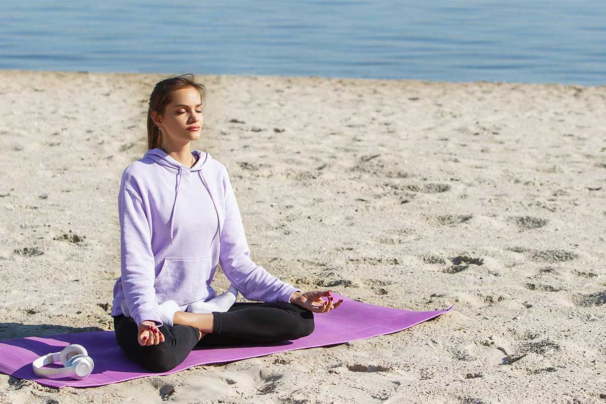 Eine junge Frau in violetten Sportklamotten sitzt am Strand und macht Yoga im Ostseeurlaub bei Private Palace Hotels & Resorts.